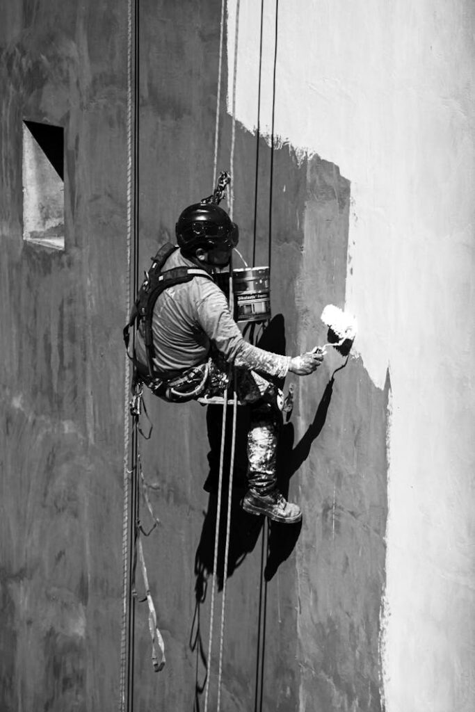 Skilled worker in action painting a tall building facade using rope access techniques.