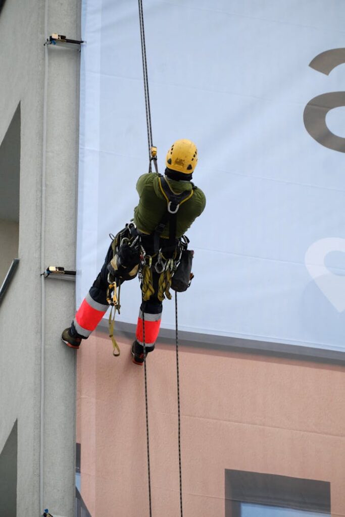 Worker rappelling down building to install large advertisement poster, wearing safety gear.